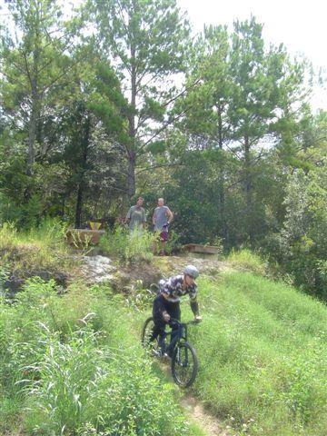 A young person rides a bicycle down a grassy hill, surrounded by trees. Two individuals stand in the background, observing the scene. The area appears to be a natural outdoor setting, possibly a bike trail or park. Santos mountain bike trail.