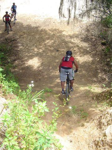 A group of mountain bikers navigating a dirt trail surrounded by greenery. The focus is on one rider in a red shirt and gray shorts, who is biking down a sloped path. Two other riders are visible in the background. The scene is sunny and showcases a natural, rugged landscape. Santos mountain bike trail.
