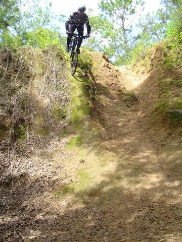 A mountain biker in a black and camouflage outfit executes a jump over a dirt trail, surrounded by greenery and trees. The bike is airborne as it navigates a steep, rugged terrain with a natural incline. Santos mountain bike trail.