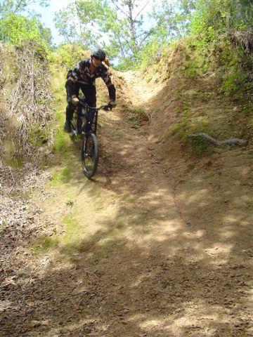 A mountain biker descending a steep, dirt trail surrounded by greenery and trees. The rider is wearing a camouflage outfit and appears focused as they navigate the incline. Santos mountain bike trail.