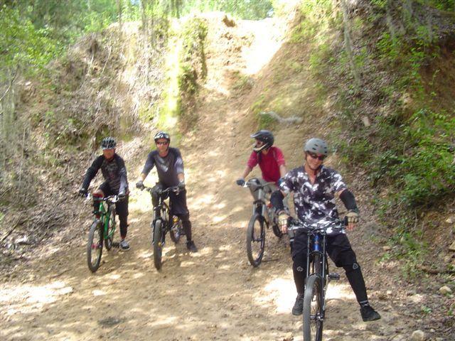 Four mountain bikers are gathered on a dirt trail surrounded by greenery. They are dressed in biking gear, including helmets, and are standing next to their bikes, which vary in color. The trail rises steeply in the background, indicating a challenging route ahead. Santos mountain bike trail.