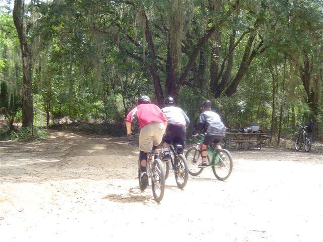 Three people on bicycles riding down a dirt path in a wooded area. The scene includes lush green trees in the background and a picnic table visible to the side. The riders are wearing helmets and protective gear as they navigate the trail. Santos mountain bike trail.