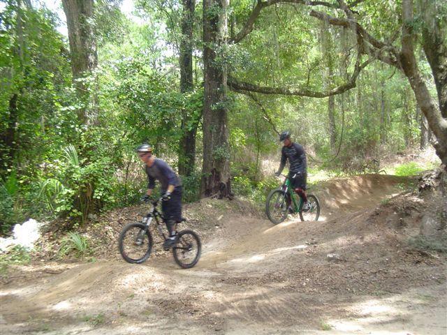 Two mountain bikers maneuvering on a dirt trail through a lush green forest. The riders are wearing athletic clothing and helmets, with one bike slightly airborne as they navigate a small ramp. Sunlight filters through the trees, creating a vibrant outdoor scene. Santos mountain bike trail.