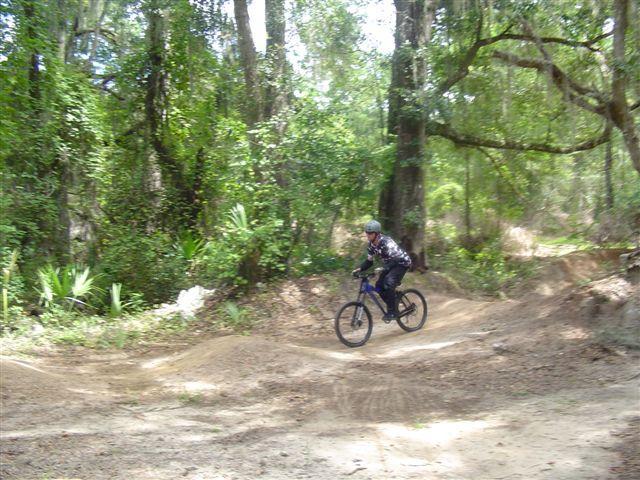 A person riding a blue bicycle on a dirt trail through a wooded area, surrounded by green foliage and trees. The rider is wearing a helmet and a patterned jacket, navigating a slight incline on the path. Santos mountain bike trail.