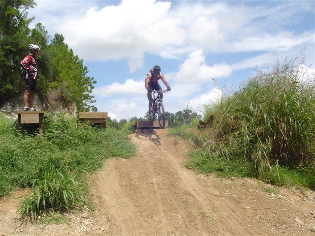 A mountain biker performs a jump off a ramp on a dirt trail, with a second cyclist watching from a nearby platform. The scene is set outdoors, featuring lush greenery and a partly cloudy sky. Santos mountain bike trail.
