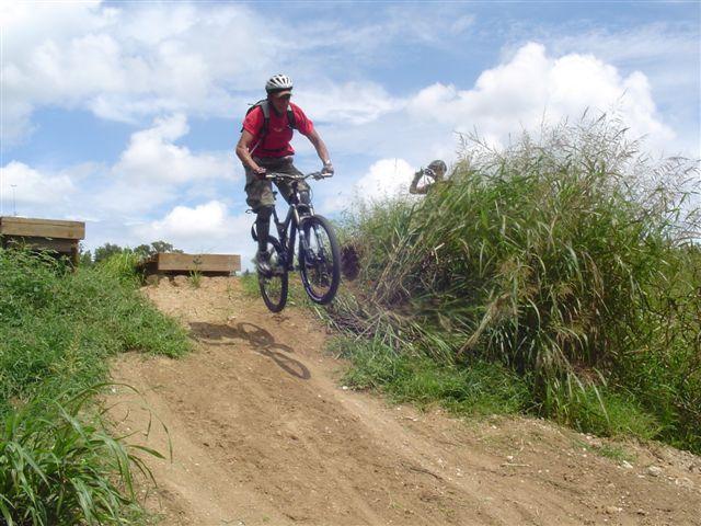 A person in a red shirt and helmet is mid-air while riding a mountain bike over a dirt ramp, surrounded by tall grass and under a blue sky with clouds. Another individual is visible in the background, observing the action. Santos mountain bike trail.