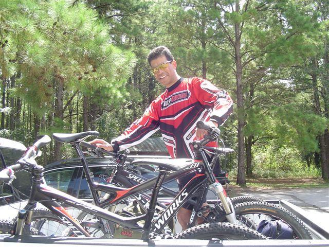 A smiling person wearing a red and black cycling jersey poses next to two mountain bikes on a truck bed, surrounded by tall pine trees in a sunny outdoor setting. Santos mountain bike trail.