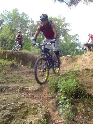 A mountain biker navigating a rocky downhill trail, wearing a helmet and protective gear. Two other cyclists can be seen in the background, surrounded by greenery. Santos mountain bike trail.