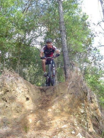 A mountain biker navigating a rocky path on a bike, surrounded by trees in a wooded area. The cyclist is leaning forward, preparing to descend a steep incline. Santos mountain bike trail.