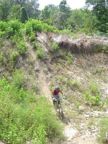 A mountain biker navigating a rugged path surrounded by lush greenery and overgrown vegetation. The trail is sloped and uneven, with the biker focused on maintaining balance while riding downhill. Santos mountain bike trail.