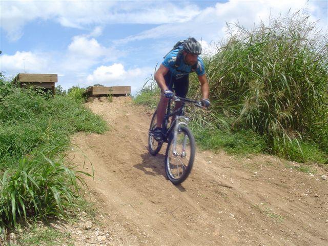 A cyclist in a blue shirt descends a dirt slope on a mountain bike, surrounded by tall grass and under a partly cloudy sky. Santos mountain bike trail.