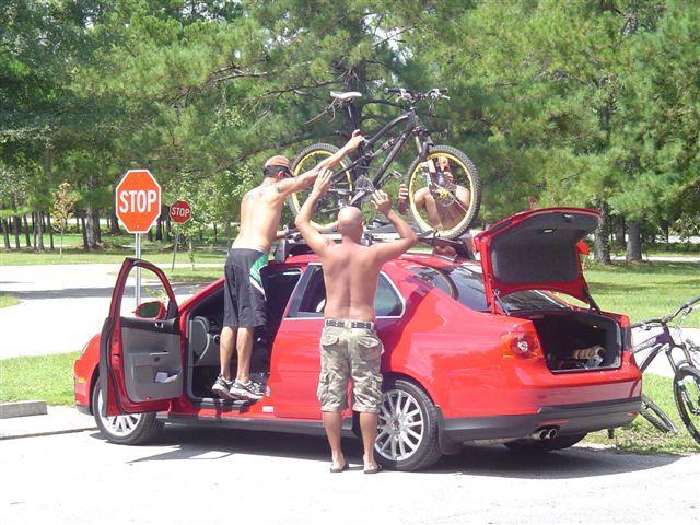 Two shirtless men are loading a bicycle onto the roof of a red car parked near a stop sign in a green, wooded area. One man stands on the driver's side of the car, lifting the bike, while the other supports it from the side. The car's doors are open, and there are additional bicycles leaning against the vehicle. Santos mountain bike trail.