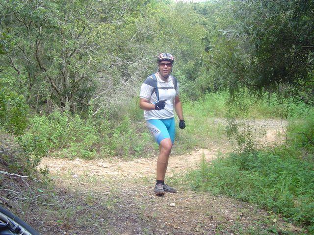 A cyclist in a light-colored shirt and blue shorts stands on a dirt path surrounded by greenery and trees. He is wearing a helmet and gloves, giving a thumbs-up gesture while looking at the camera. Santos mountain bike trail.