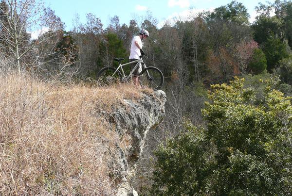 A person in a white helmet stands on the edge of a rocky cliff while holding a mountain bike, overlooking a wooded area below. The landscape features dry grass and trees in various shades of green and brown, with a clear blue sky above. Santos mountain bike trail.
