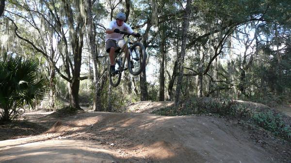 A mountain biker performing a jump on a dirt ramp amidst a wooded area with trees and vegetation in the background. The rider is wearing a helmet and casual clothing, showcasing an action-packed moment in a natural setting. Santos mountain bike trail.