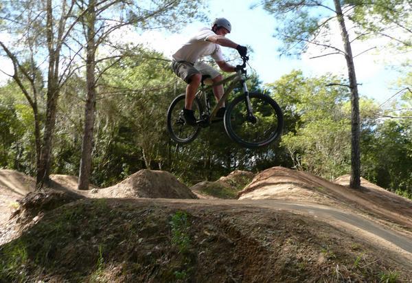 A cyclist performs a jump on a mountain bike over a dirt ramp in a forested area, surrounded by trees and dirt trails. Santos mountain bike trail.