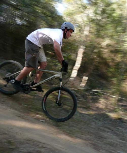 A person riding a mountain bike on a dirt trail surrounded by trees, captured in a dynamic, blurred motion that conveys speed and excitement. The rider is wearing a helmet and casual athletic clothing, with a focus on their active position as they navigate the terrain. Santos mountain bike trail.