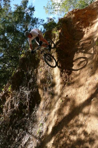 A mountain biker executes a downhill maneuver on a steep, dirt trail surrounded by trees. The rider is wearing a helmet and is positioned low on the bike, capturing a dynamic moment in action. Sunlight filters through the foliage, creating a mix of shadows and highlights on the trail. Santos mountain bike trail.