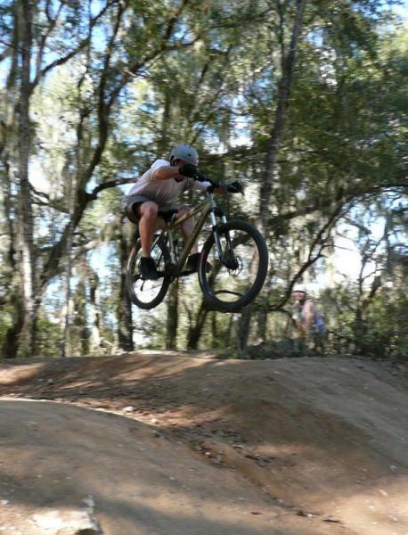 A person in a helmet performs a jump on a mountain bike over a dirt ramp, surrounded by trees and natural foliage. Santos mountain bike trail.