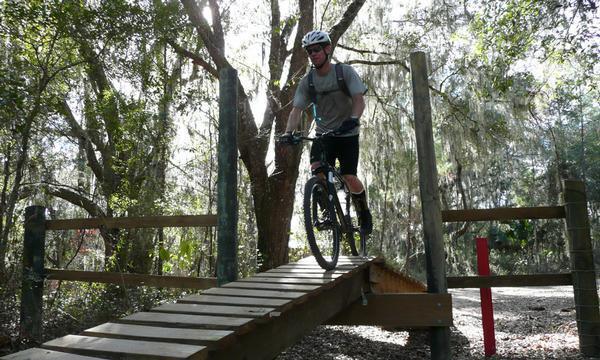 A cyclist riding a mountain bike over a wooden bridge in a forested area, surrounded by trees and dappled sunlight. The rider is wearing a helmet and cycling attire, showcasing a dynamic moment of biking on a trail. Santos mountain bike trail.