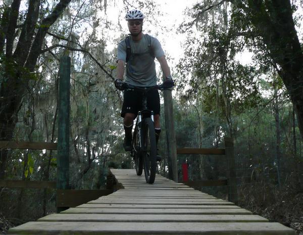 A cyclist riding a mountain bike over a wooden bridge in a forested area, surrounded by trees and foliage. The rider is wearing a helmet and athletic clothing, navigating the narrow path with concentration. Santos mountain bike trail.
