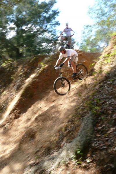 A mountain biker in a helmet rides down a steep, dirt slope, capturing a dynamic moment of descent. In the background, another person watches from a higher vantage point among trees. Santos mountain bike trail.