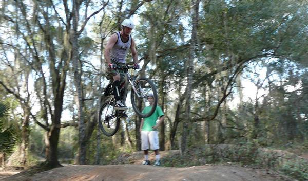 A cyclist in mid-air performing a jump on a mountain bike, surrounded by trees in a forested area. A second person stands nearby, watching the action. The cyclist is wearing a helmet and tank top, while the onlooker is dressed in a green shirt and white shorts. Santos mountain bike trail.