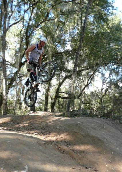 A mountain biker performing a jump on a dirt ramp in a wooded area, surrounded by trees with green foliage. The rider is wearing a helmet and a sleeveless shirt, showcasing a dynamic action shot. Santos mountain bike trail.