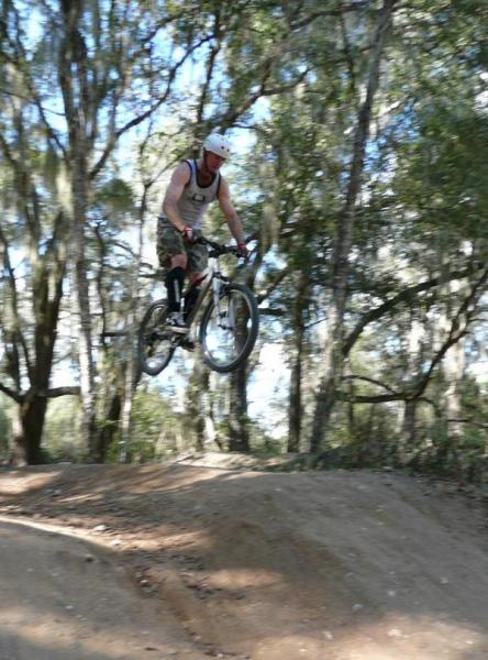 A person in a tank top and helmet is performing a jump on a mountain bike, airborne above a dirt ramp surrounded by trees. Santos mountain bike trail.