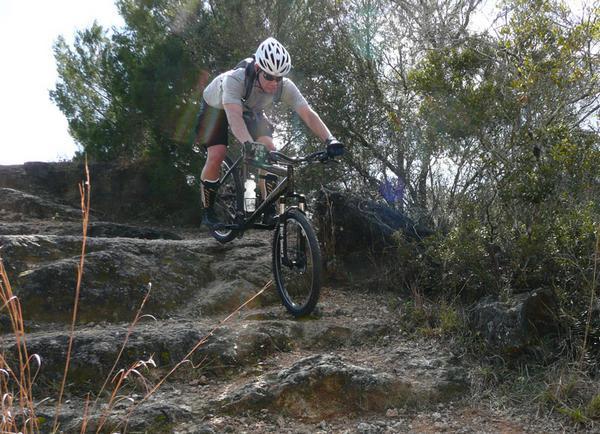 A mountain biker expertly navigating rocky terrain, captured in mid-air above the rocks. The rider wears a helmet and protective gear, demonstrating an adventurous spirit in a natural environment filled with shrubs and trees. Santos mountain bike trail.