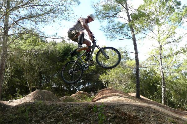A person performing a jump on a mountain bike over a dirt ramp, surrounded by trees and a clear blue sky. Santos mountain bike trail.