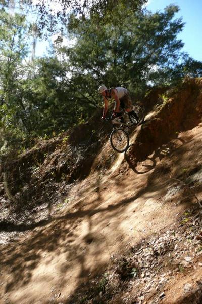 A mountain biker launches off a dirt ramp in a wooded area, with green trees in the background and a clear blue sky above. Santos mountain bike trail.