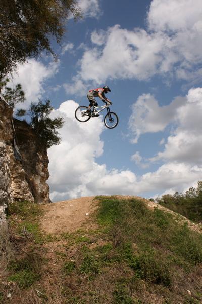 A mountain biker performing a jump over a dirt ramp, soaring above a rocky cliff against a backdrop of blue sky with fluffy white clouds. Santos mountain bike trail.