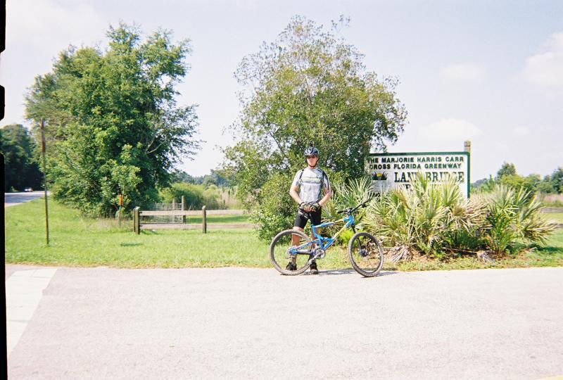 A cyclist wearing a helmet and a sporty outfit stands next to a blue mountain bike at the Marjorie Harris Carr Cross Florida Greenway sign. The background features lush greenery and a sunny sky, creating a vibrant outdoor scene. Santos mountain bike trail.