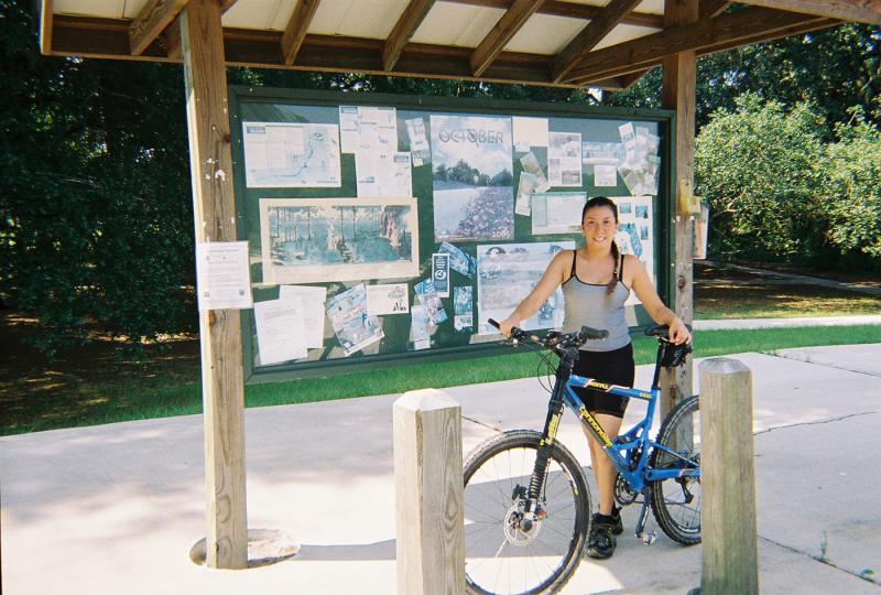 A young woman stands next to a blue mountain bike in front of an outdoor information board covered with maps and notices. She is wearing a sleeveless gray top and black shorts, smiling at the camera. The setting is a park with greenery visible in the background. Santos mountain bike trail.