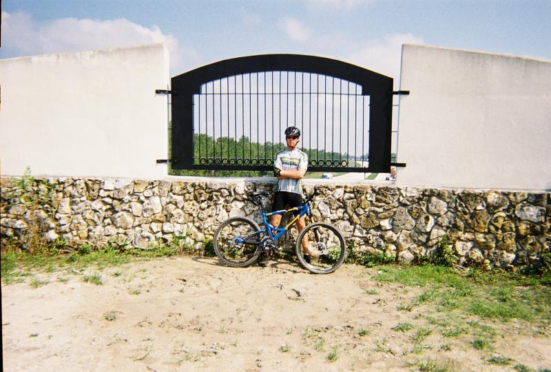 A person in a cycling outfit stands beside a blue mountain bike, leaning against a stone wall. Behind them is a gated entrance with a decorative arch, set against a clear sky with a few clouds. The surrounding area features sandy ground and patches of grass, suggesting a rural or outdoor setting. Santos mountain bike trail.