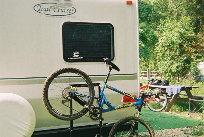 A blue mountain bike is mounted on a stand next to a travel trailer labeled "Trail Cruiser." In the background, there are picnic tables and trees, creating a campsite atmosphere. Some outdoor gear is visible on a nearby table. Santos mountain bike trail.