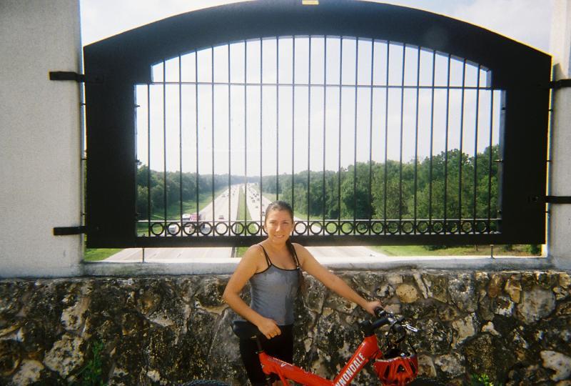 A young woman standing next to a red bicycle, smiling at the camera. She is positioned in front of a decorative gate overlooking a highway and lush green trees in the background. The sky is bright and sunny. Santos mountain bike trail.