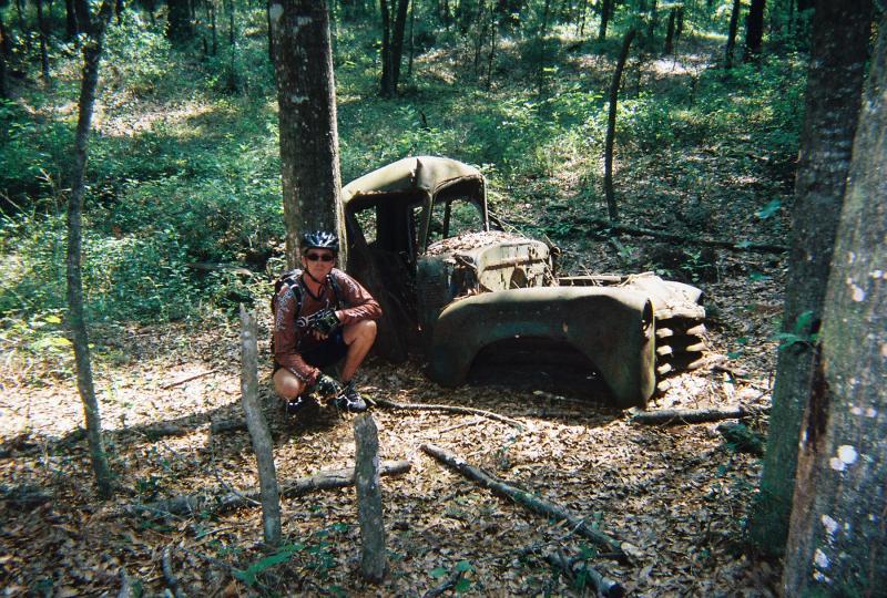 A person wearing a bicycle helmet crouches next to an old, rusty truck abandoned in a wooded area, surrounded by trees and fallen leaves. Santos mountain bike trail.