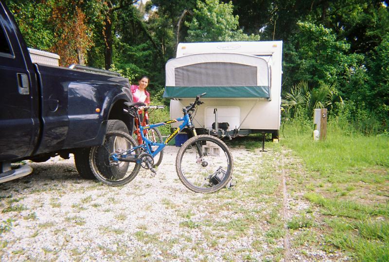 A blue mountain bike is parked next to a black truck, with a camper trailer in the background. A woman is smiling while standing beside the bike, surrounded by lush greenery and gravel. A wooden post marked with the number "5" is visible to the right. Santos mountain bike trail.