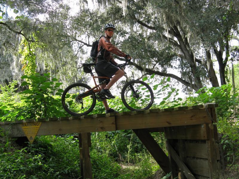 A cyclist wearing a helmet and a backpack balances on a wooden bridge in a lush, green forest. Sunlight filters through the trees above, and there are various plants surrounding the area. A warning sign is visible on the bridge. Santos mountain bike trail.