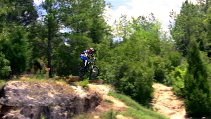 A mountain biker performing a jump off a rocky ledge, surrounded by lush green trees under a clear blue sky. Santos mountain bike trail.