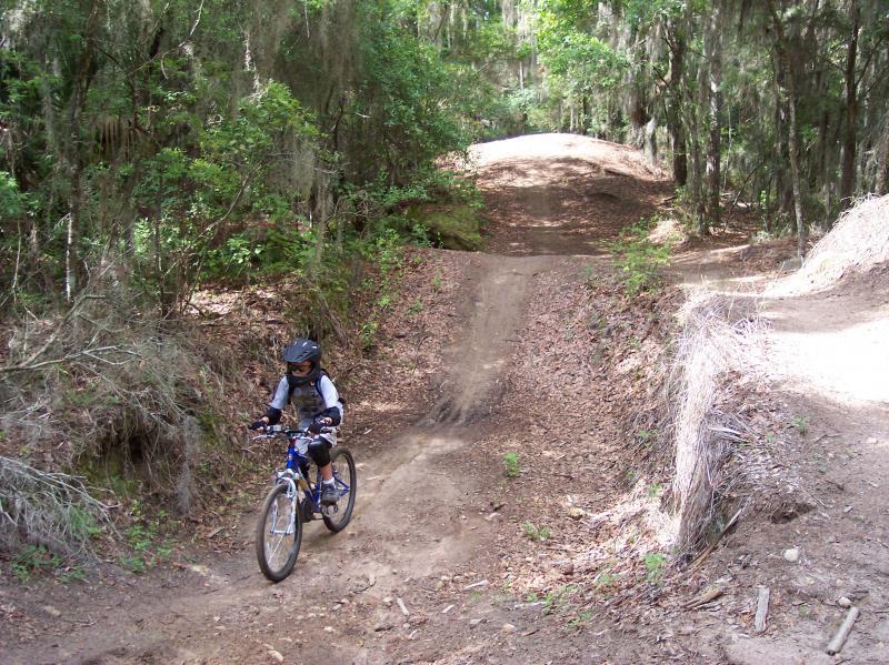 A child in protective gear rides a mountain bike on a dirt trail in a wooded area, navigating a small hill. The surroundings feature dense greenery and scattered leaves, creating an outdoor biking environment. Santos mountain bike trail.
