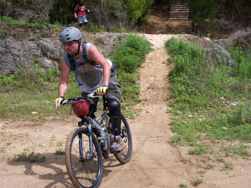 A man riding a mountain bike on a dirt trail, wearing a helmet, knee pads, and gloves. In the background, another person is walking up a hill towards a wooden structure. The surrounding area is lush with greenery and rocky terrain. Santos mountain bike trail.