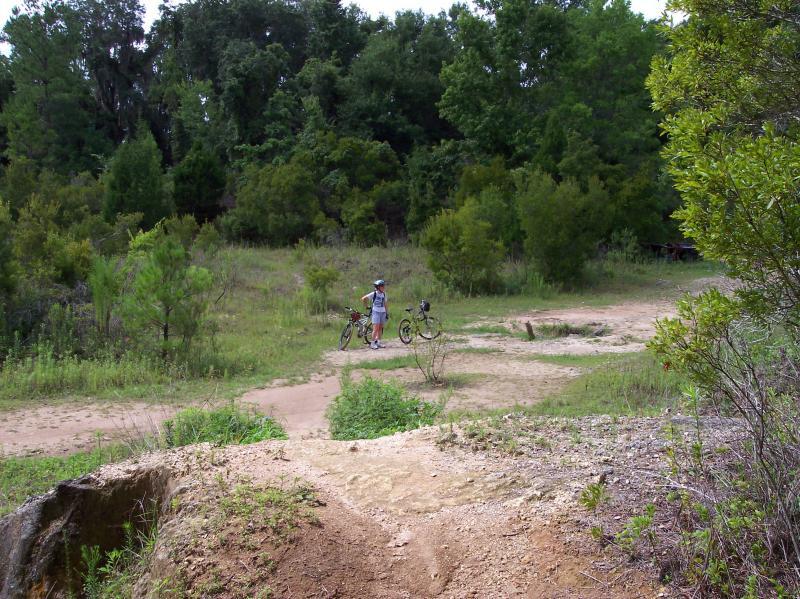 A person stands beside two bicycles on a dirt trail surrounded by greenery. The scene features a hilly terrain with multiple bike paths converging, set against a backdrop of lush trees and bushes under a clear sky. Santos mountain bike trail.