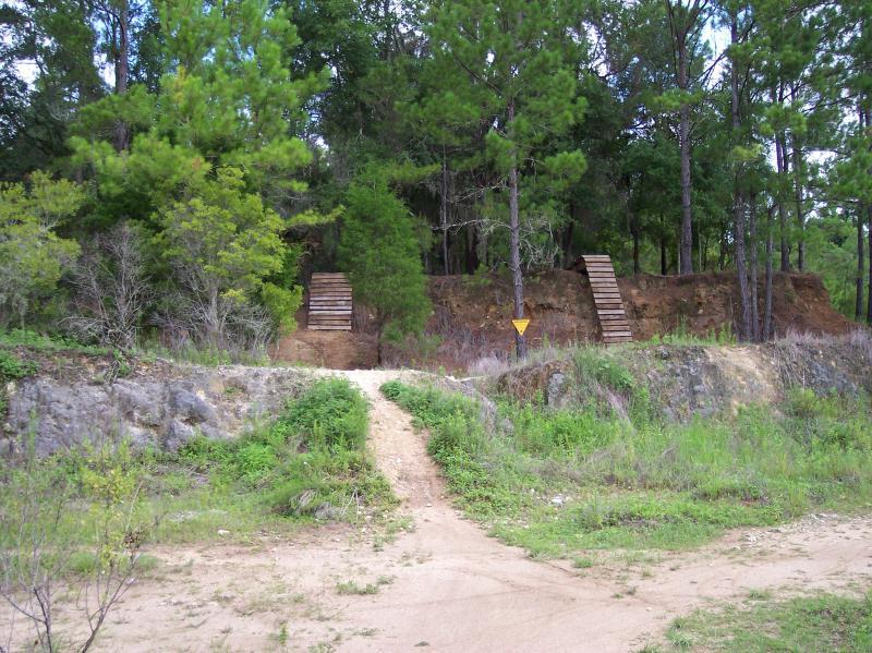 A sandy pathway leads to a slope with wooden steps on either side, surrounded by dense greenery and tall trees. A caution sign is positioned near the top of the slope, indicating potential hazards. Santos mountain bike trail.