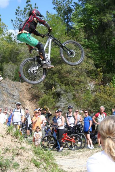 A mountain biker performing a trick jump over a dirt ramp, while a crowd of spectators watches nearby, enjoying the outdoor event in a lush, green environment. Santos mountain bike trail.