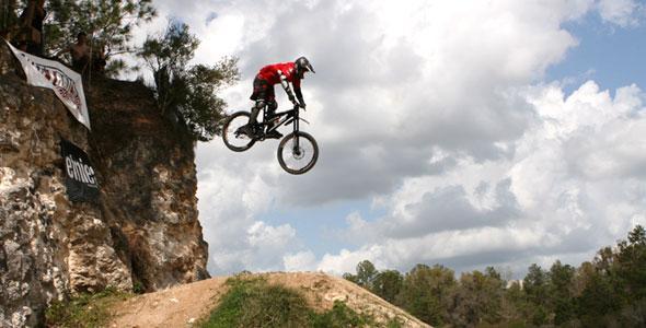 A mountain biker in a red jersey and black helmet performing a jump off a rocky ledge, with clouds and trees in the background. Santos mountain bike trail.