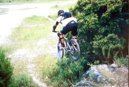 A mountain biker is mid-air, leaping off a rock ledge, with greenery and a dirt path visible in the background. The cyclist is wearing a helmet, a black backpack, and protective gear. Santos mountain bike trail.