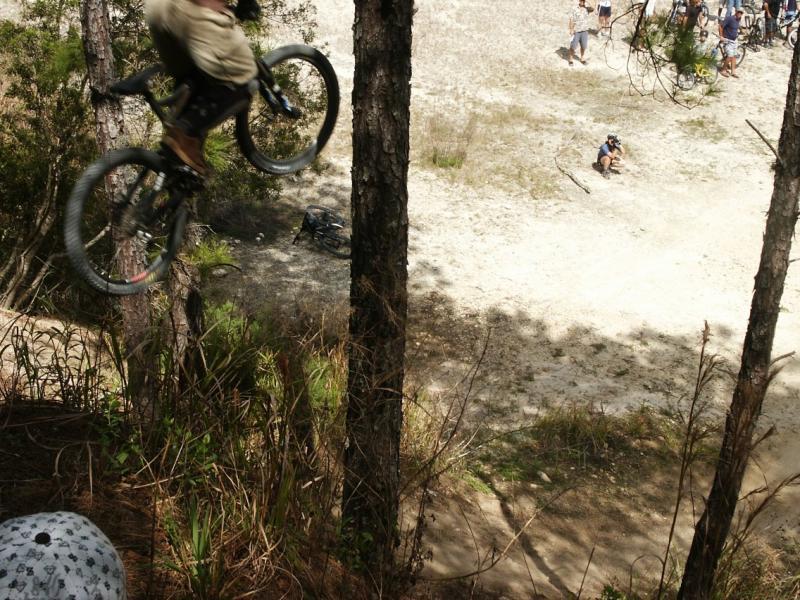 A cyclist performing a jump over a dirt ramp in a wooded area, with trees in the foreground and other riders in the background. Santos mountain bike trail.